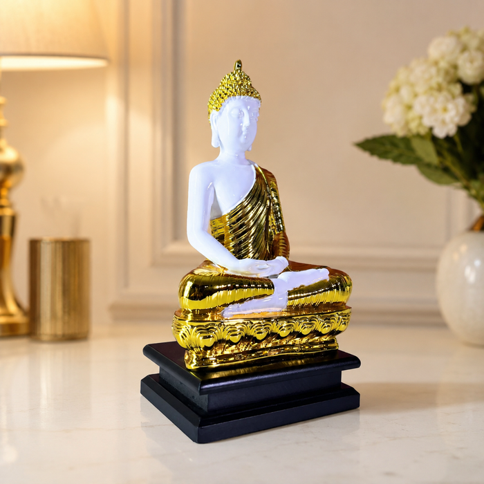 A white and gold Buddha statue seated in a meditative pose on a black base, displayed on a tabletop with softly blurred home decor in the background.