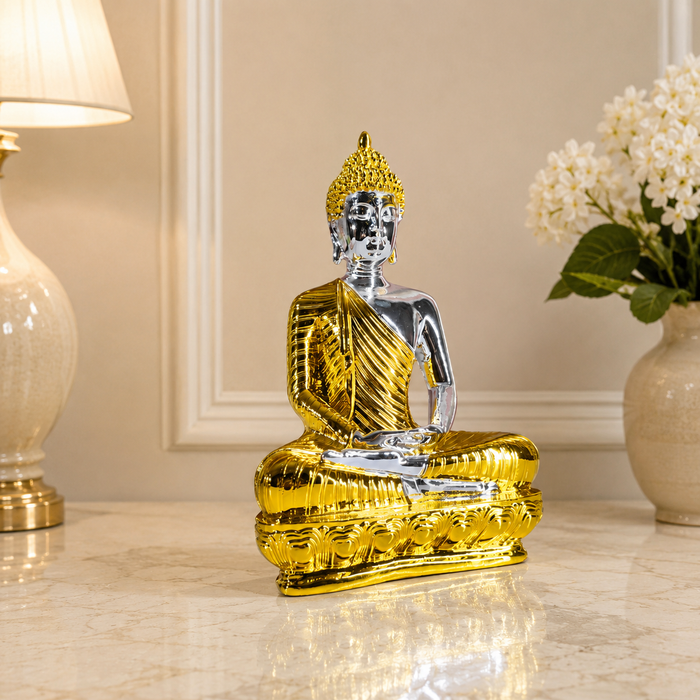 A metallic Buddha statue in a seated meditation pose, featuring a shiny gold finish on the robe and base with a polished silver finish on the face and arms, displayed on a marble tabletop with a lamp and flowers in the background.