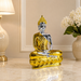 A metallic Buddha statue in a seated meditation pose, featuring a shiny gold finish on the robe and base with a polished silver finish on the face and arms, displayed on a marble tabletop with a lamp and flowers in the background.