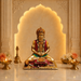 A gold-toned seated deity statue with red and green draped details, placed on a marble surface in front of a warm-lit arched alcove. Decorative flowers, brass oil lamps, and a small brass object sit on both sides.
