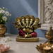 A gold-toned multi-headed Ganesha statue with red drapery sits on a tabletop in a styled interior setting with flowers, a brass pot, and lit candles.