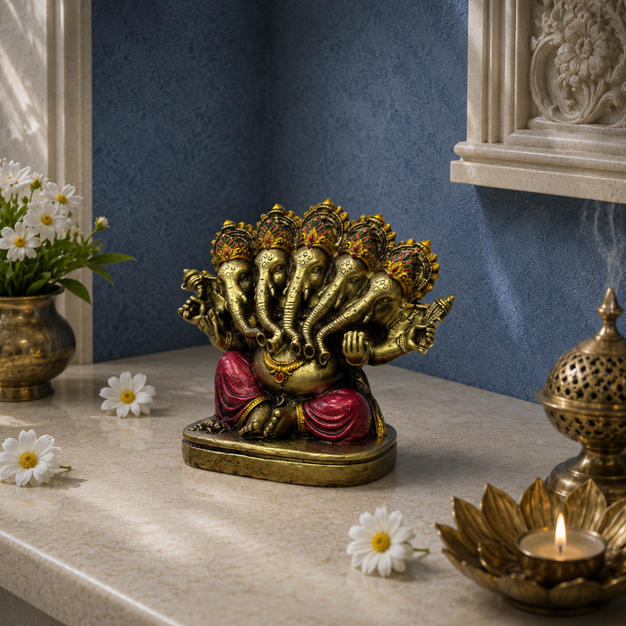 A gold-toned multi-headed Ganesha statue with red draped fabric, displayed on a beige marble tabletop in a styled interior setting with blue walls, white daisies, a brass pot, and a lit brass incense burner.