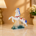 A decorative porcelain horse figurine with a white body, blue mane and tail, red harness details, and a castle-and-water base, displayed on a beige tabletop with a lamp and flowers in the background.