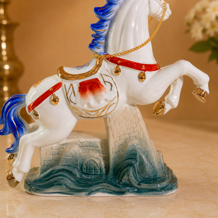 A decorative porcelain horse figurine with a white body, blue mane and tail, red harness details, gold trim, and a blue-gray wave base, shown on a warm beige surface.
