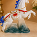 A decorative porcelain horse figurine with a white body, blue mane and tail, red harness details, gold trim, and a blue-gray wave base, shown on a warm beige surface.