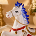 A glossy ceramic rocking horse figurine with a white body, vibrant blue mane, red saddle blanket, and gold decorative accents, shown in a warm indoor setting.