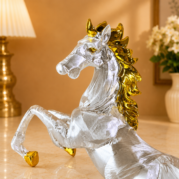 A decorative glass horse figurine with a transparent body and shiny gold accents on the mane and hooves, displayed on a tabletop in a warmly lit indoor setting.