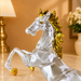 A decorative glass horse figurine with a transparent body and shiny gold accents on the mane and hooves, displayed on a tabletop in a warmly lit indoor setting.