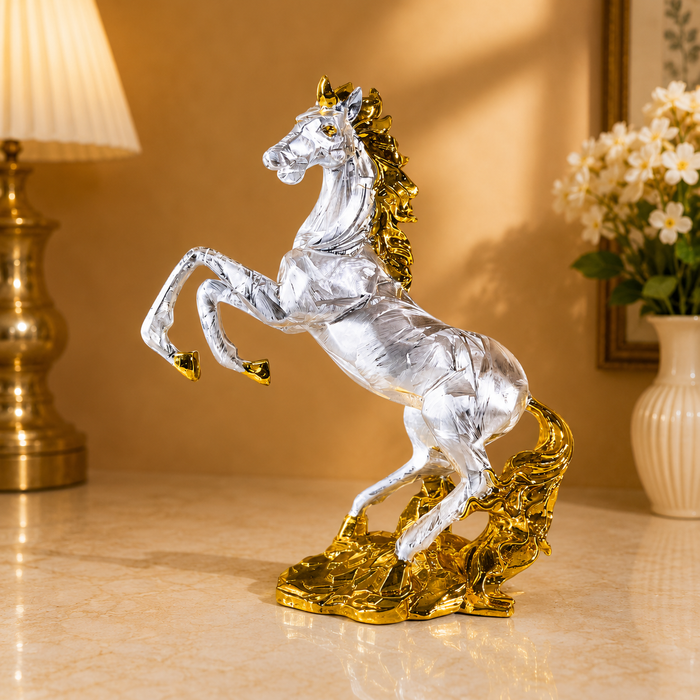 A metallic silver horse figurine with a gold mane, hooves, and base, shown on a polished marble surface in a warmly lit room with a lamp and flowers in the background.