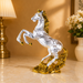 A metallic silver horse figurine with a gold mane, hooves, and base, shown on a polished marble surface in a warmly lit room with a lamp and flowers in the background.
