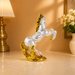 A clear glass rearing horse figurine with metallic gold accents on the mane, hooves, and base, displayed on a polished marble tabletop in a warm, elegant room setting.