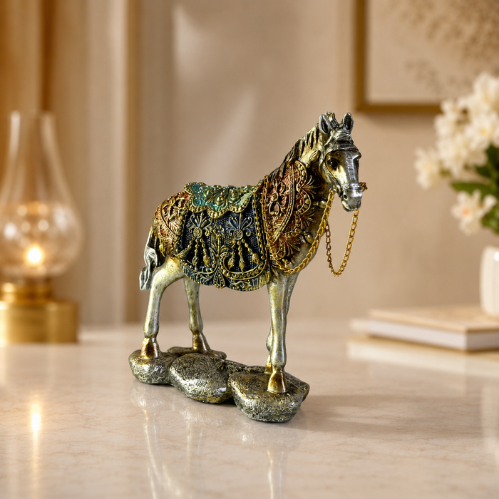 An ornate horse figurine with a metallic silver body and intricate gold, green, and blue decorative patterns on its saddle and harness, standing on a textured base on a light marble surface.