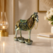 An ornate horse figurine with a metallic silver body and intricate gold, green, and blue decorative patterns on its saddle and harness, standing on a textured base on a light marble surface.