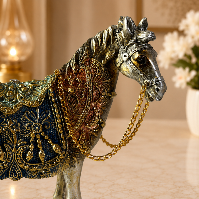 An ornate decorative horse figurine with a metallic silver body, detailed with a dark blue and gold patterned saddle blanket and a gold chain halter, set against a softly blurred indoor background.