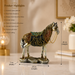 An ornate decorative horse statue with a metallic silver body and an intricately patterned saddle in green, gold, and red, standing on a textured base. The image includes product dimensions (20 cm length, 7 cm width, 24 cm height) and a list of product highlights on the left, set against a warm, softly blurred home background.