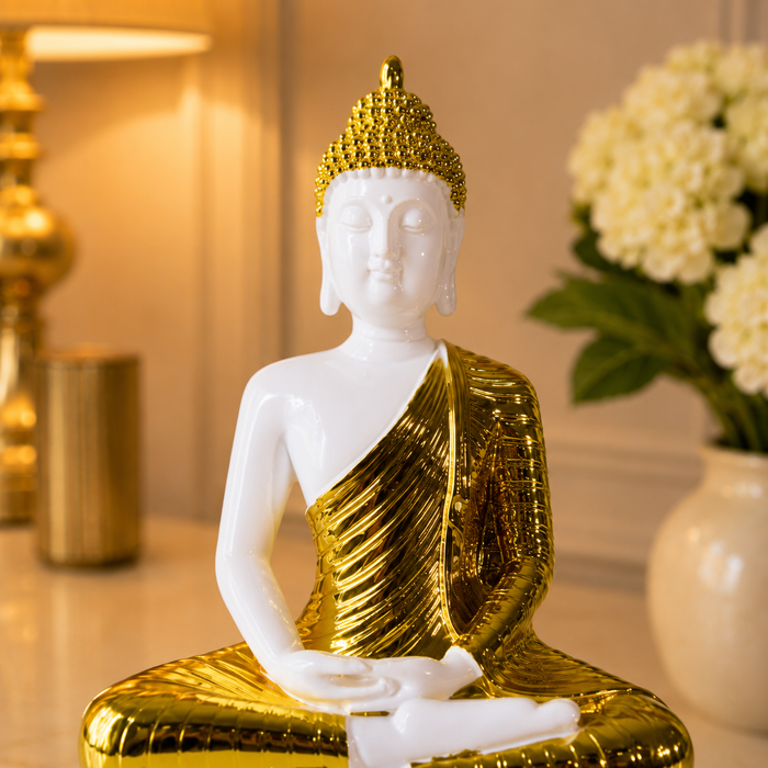 A seated Buddha figurine with a glossy white body and a metallic gold robe and crown, displayed on a tabletop with warm indoor lighting and blurred flowers in the background.