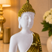 A glossy white Buddha statue with a textured gold crown and draped gold robe, shown in a warmly lit indoor setting with a lamp and flowers blurred in the background.