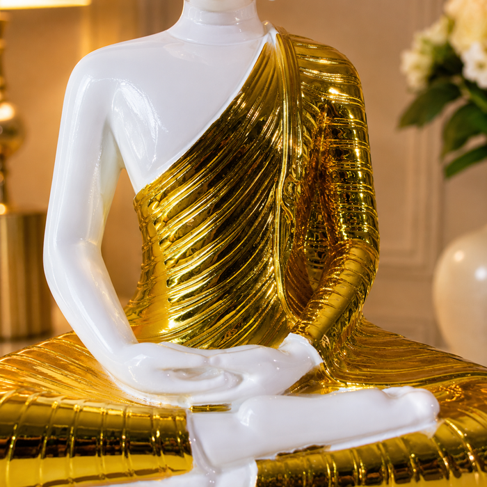 A seated Buddha statue with a glossy white body and a metallic gold draped robe, shown in a warmly lit indoor setting with blurred decor in the background.