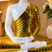 A seated Buddha statue with a glossy white body and a metallic gold draped robe, shown in a warmly lit indoor setting with blurred decor in the background.