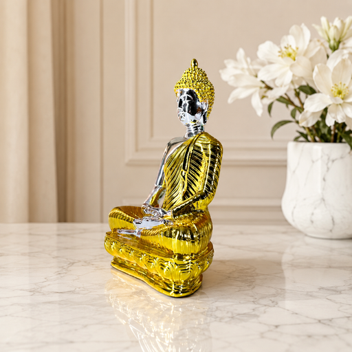 A metallic gold Buddha statue in a seated meditation pose, displayed on a light marble surface with a vase of white flowers in the background.