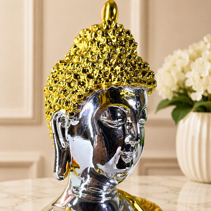 A decorative Buddha head sculpture with a highly reflective silver-toned face and a textured, metallic gold hairpiece, set on a light marble surface with a blurred background of white flowers.