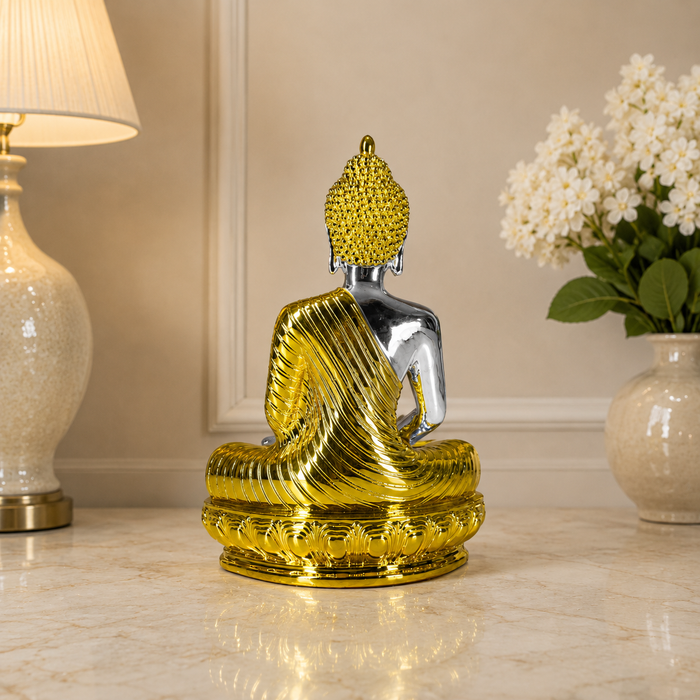 A decorative seated Buddha statue shown from the back, with a metallic gold textured robe and head, a polished silver-toned body, and a gold lotus-style base. The statue is placed on a light marble surface in an indoor setting with a lamp on the left and white flowers in a vase on the right.