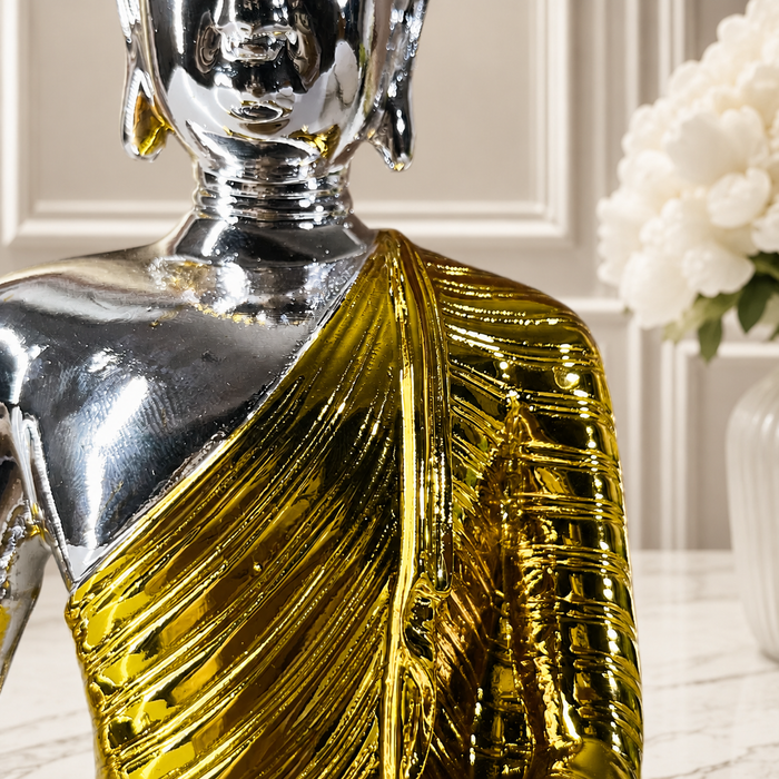 A decorative Buddha statue with a highly reflective silver metallic finish on the head and shoulders, and a textured, ribbed gold draped robe, shown on a light marble surface with a blurred white floral background.