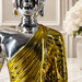 A decorative Buddha statue with a highly reflective silver metallic finish on the head and shoulders, and a textured, ribbed gold draped robe, shown on a light marble surface with a blurred white floral background.
