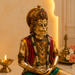 A golden Hanuman statue with red and green draped fabric, adorned with detailed jewelry, set against a warm beige background with a lit candle and soft architectural details.