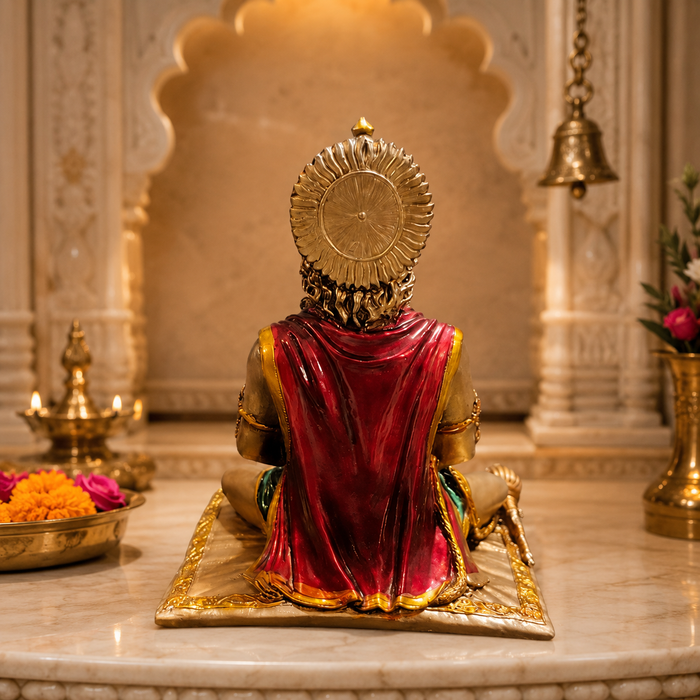 A golden statue of a seated Hindu deity, viewed from behind, draped in a rich red shawl, placed on a marble altar in a temple setting with brass oil lamps and flowers.