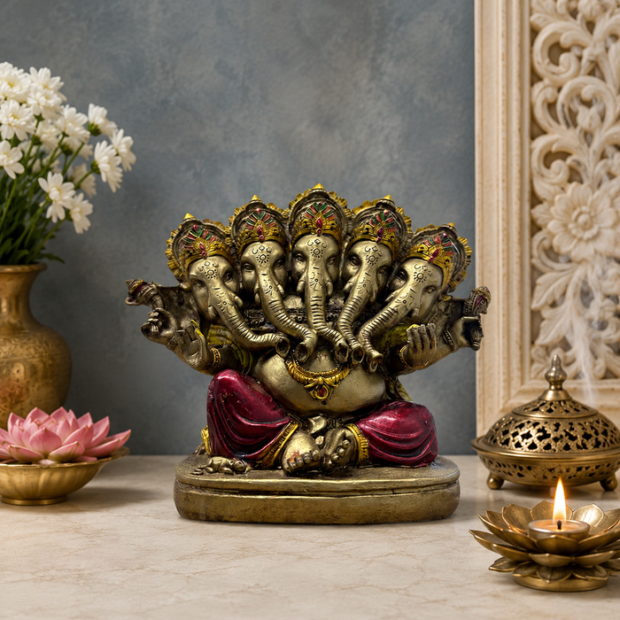 A brass Ganesha idol with multiple elephant heads and a red draped skirt, displayed on a marble tabletop with decorative props including a lotus flower, a lit brass incense burner, and white flowers in a vase.