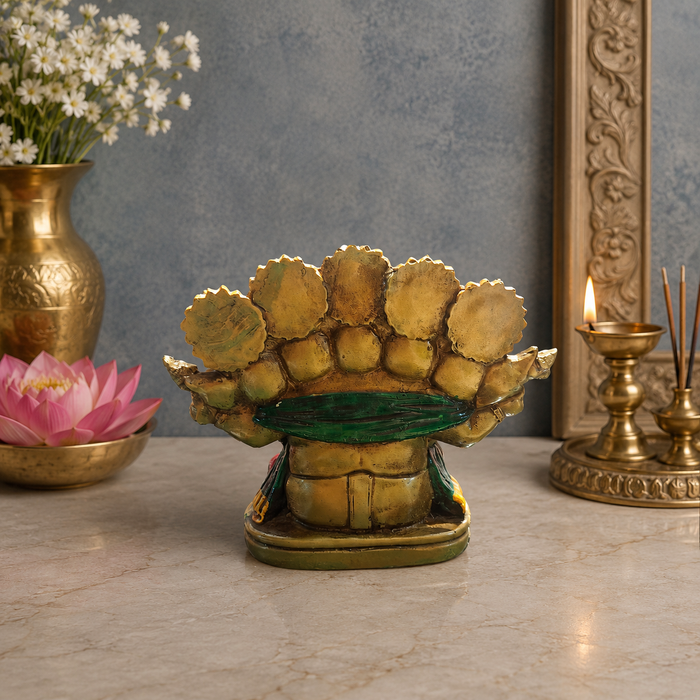 A decorative golden lotus bud sculpture or candle holder with a green band around the middle, displayed on a marble surface in a styled interior setting with a vase of white flowers, a pink lotus, and brass candlesticks in the background.