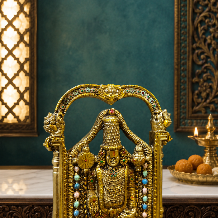 An ornate gold-toned Hindu deity statue seated on a decorated throne, set on a marble surface against a teal wall with traditional decor and a lit brass oil lamp in the background.