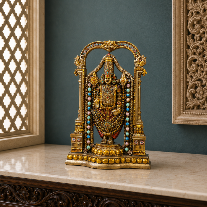 An ornate gold-toned Hindu deity statue, likely Lakshmi, displayed on a polished marble console table against a dark teal wall. The statue is decorated with intricate gold jewelry, red garments, and rows of turquoise and white beads, framed by decorative pillars.