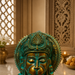 A close-up of a decorative metallic idol with a turquoise-green patina and gold-toned face, set against an ornate warm brown interior background with carved wood, a lit candle, and a vase.