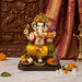 A colorful Ganesha idol seated on a decorative pedestal, with ornate carved wood in the background and small offerings placed around it on a marble surface.