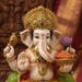 A close-up of a Ganesha statue with a cream-colored face and body, decorated with a large gold crown and jewelry, holding a mace and a lotus flower with an orange bowl. The background is a softly blurred warm brown and gold setting.