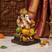 A colorful Ganesha statue seated on a dark base, styled on a marble surface with decorative offerings, a carved wooden backdrop, and draped red fabric in the background.