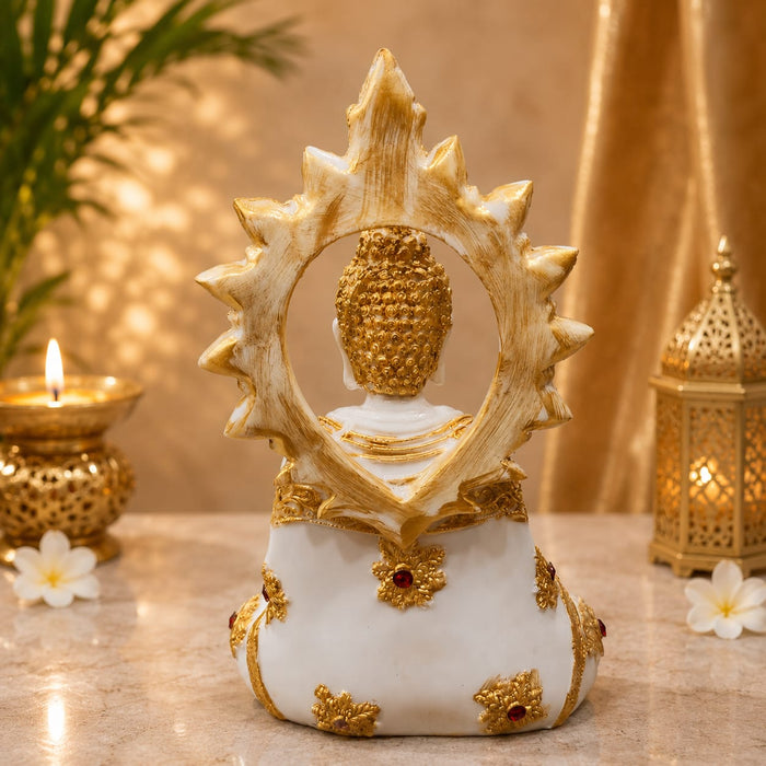 A decorative Buddha statue shown from the back, featuring a white body with gold accents and a large gold sunburst halo. The statue is displayed on a marble-like surface with a lit candle holder, a lantern, and white flowers in a warm, golden-toned setting.