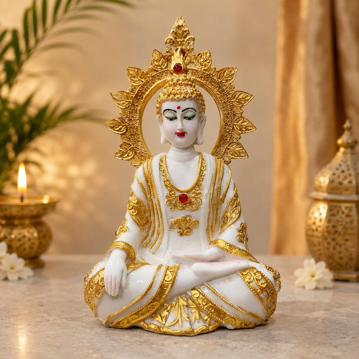 A white and gold seated Buddha statue with ornate gold halo and robe details, placed on a tabletop with warm decorative background elements including a candle, plant leaves, and a lantern.