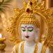 A close-up of a white and gold Buddha statue with a detailed golden halo, green eyes, and red painted features, set against a softly blurred warm background with a candle visible on the left.