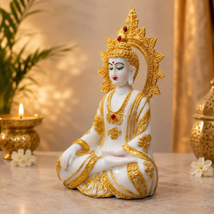 A white and gold seated Buddha statue with a detailed gold halo and crown, placed on a tabletop with decorative candles and flowers in a warm indoor setting.