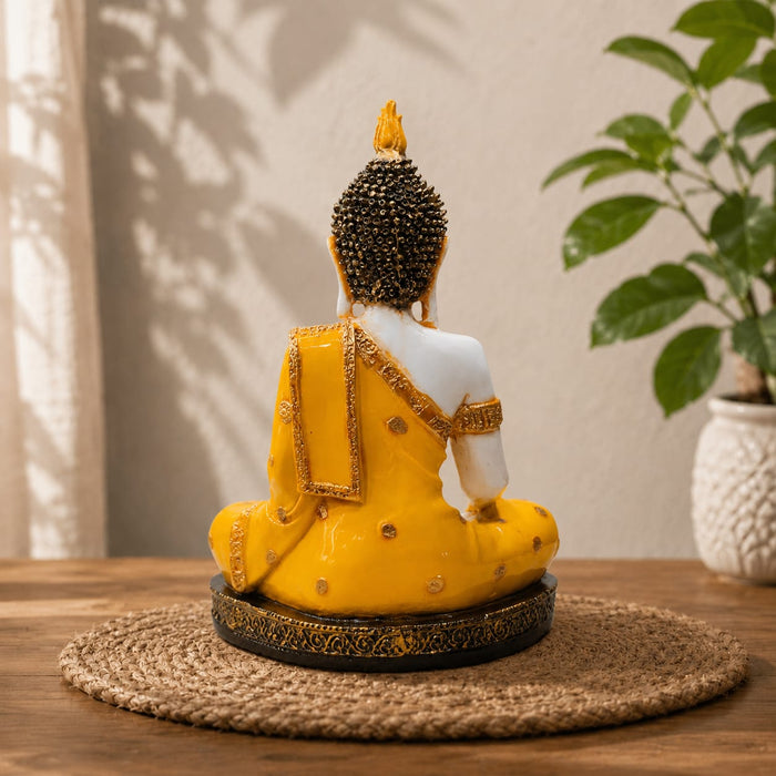 A seated Buddha figurine shown from the back, wearing a bright yellow robe with gold detailing and a white upper back, placed on a woven round mat on a wooden surface with a potted plant in the background.