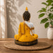 A seated Buddha figurine shown from the back, wearing a bright yellow robe with gold detailing and a white upper back, placed on a woven round mat on a wooden surface with a potted plant in the background.