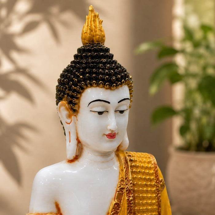 A white and gold Buddha statue shown in a close-up indoor shot, with a blurred potted plant and wall shadows in the background.