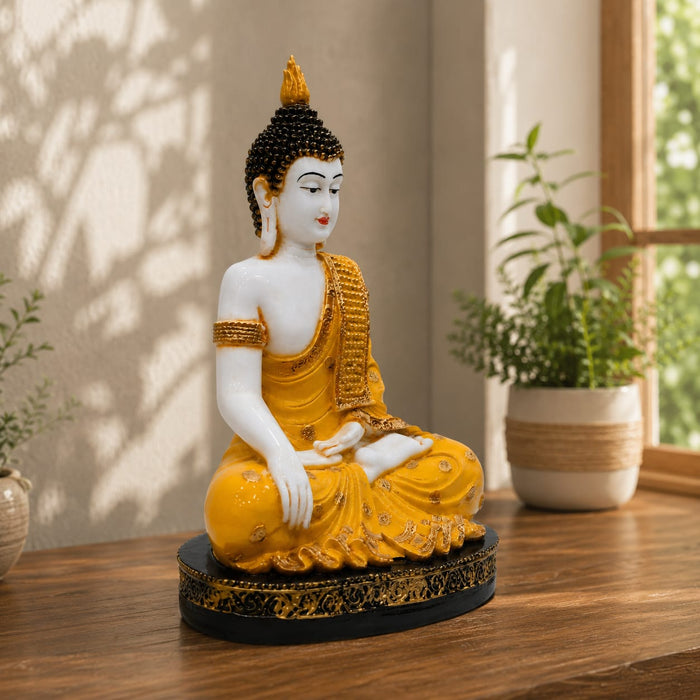 A seated Buddha statue in a meditative pose, painted in white and golden yellow with a black and gold base, displayed on a wooden table in a sunlit room with potted plants.