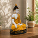 A seated Buddha statue in a meditative pose, painted in white and golden yellow with a black and gold base, displayed on a wooden table in a sunlit room with potted plants.