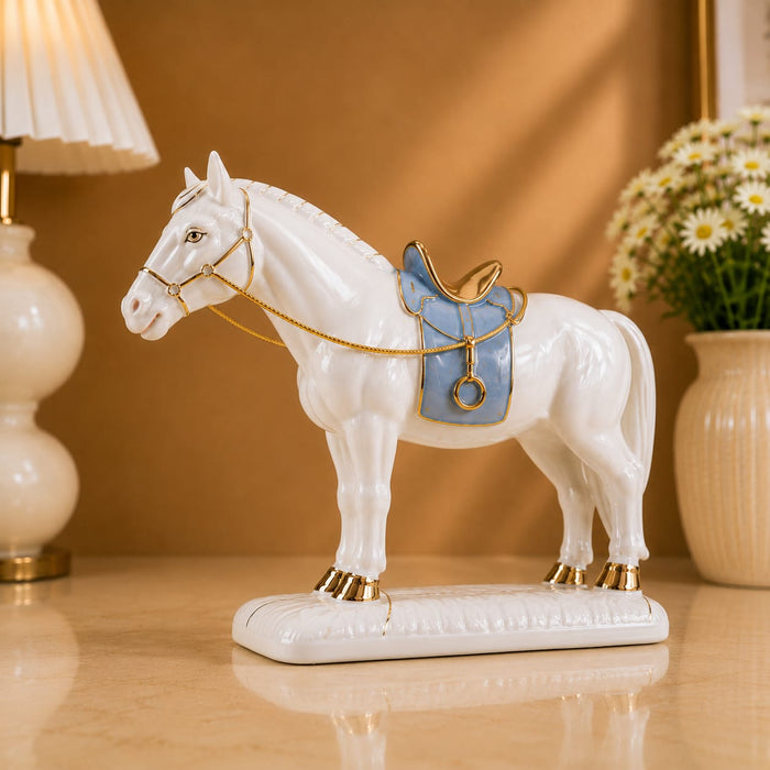 A white porcelain horse figurine with a light blue saddle and gold trim, displayed on a beige tabletop with a lamp and flowers in the background.