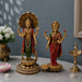 Two ornate gold-toned Hindu deity statues displayed on a tabletop against a muted blue-gray wall. The larger statue on the left depicts a multi-armed deity with a large shell canopy and red-and-green draped attire, while the smaller statue on the right shows a similarly detailed deity in red and green clothing.