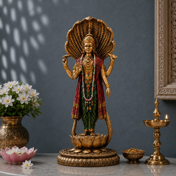 A golden Lord Shiva statue with a large open shell canopy, red and green draped garments, standing on a lotus base. The statue is displayed on a marble surface with a brass lamp, flowers, and a decorative frame against a dark gray wall.